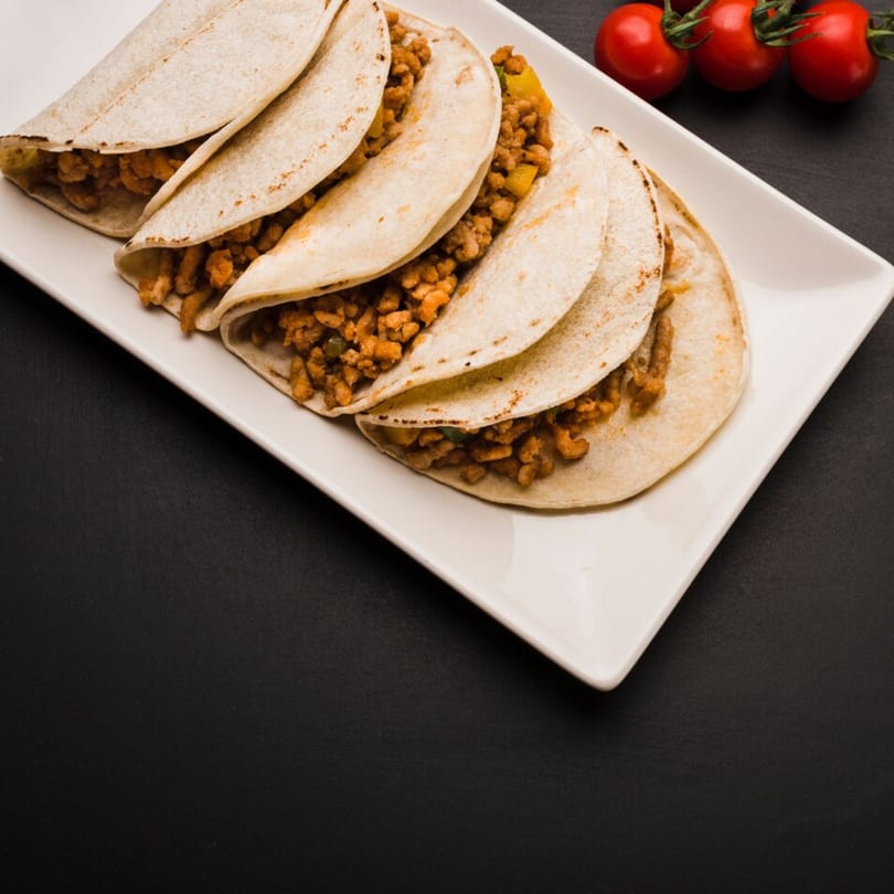 A white rectangular plate holds five tacos filled with ground meat and vegetables, with cherry tomatoes on the vine.