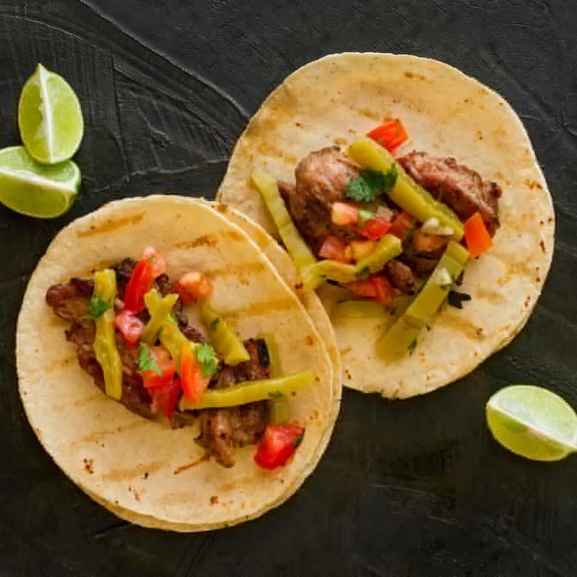 Two carne asada tacos with pico de gallo and green chiles, garnished with lime wedges.