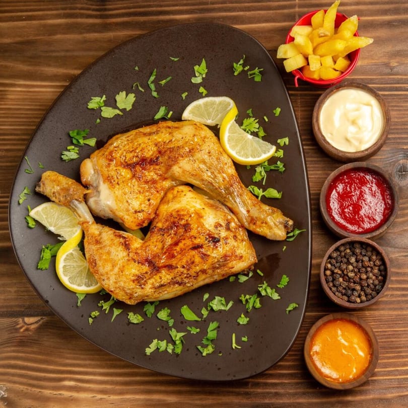 Two golden-brown roasted chicken legs with lemon slices and parsley on a dark plate, with fries and sauces.