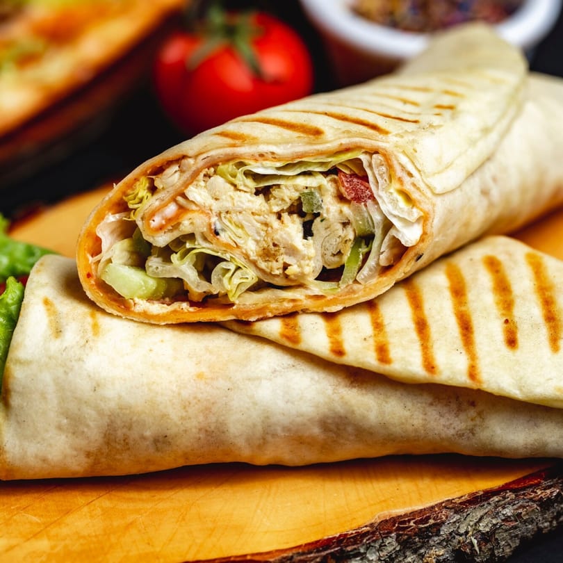 Two grilled chicken wraps with lettuce and sauce, on a wooden board with a tomato in the background.
