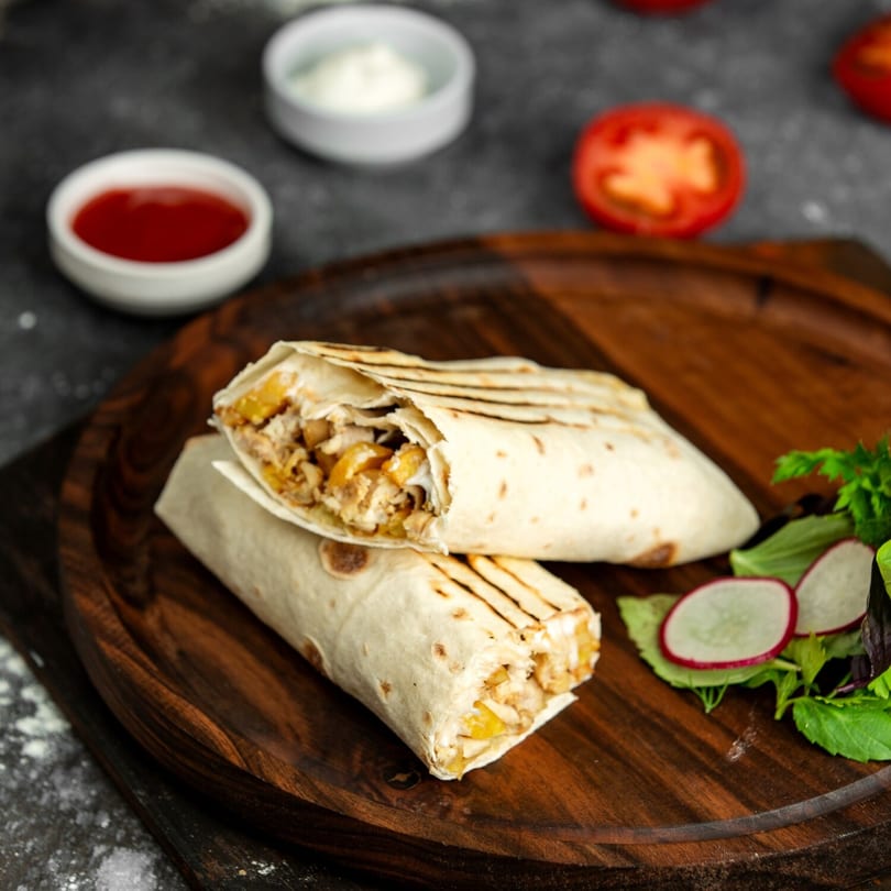 Two chicken shawarma wraps with fries, salad, and dipping sauces on a wooden board.