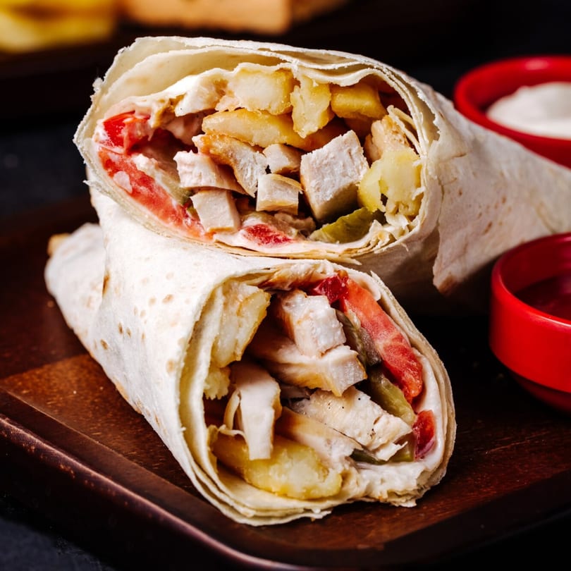 Two chicken shawarmas, filled with chicken, fries, tomatoes, and pickles, on a wooden board.