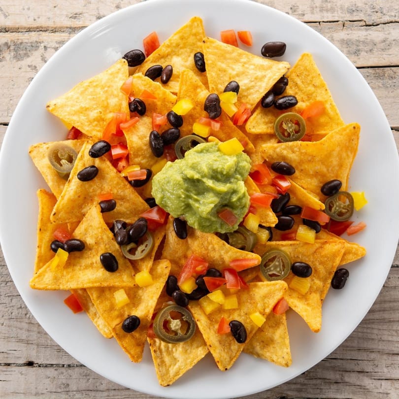 Nachos with guacamole, black beans, diced tomatoes, yellow peppers, and jalapeños on a white plate.